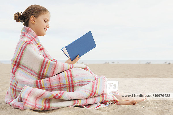 In eine Decke eingewickeltes Mädchen sitzt am Strand und liest ein Buch; Toronto  Ontario  Kanada'.