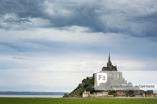 Große steinerne Abtei in der Ferne auf einer felsigen Insel mit einem grasbewachsenen Feld im Vordergrund und einer Wolkendecke; Mont St. Michel  Bretagne  Frankreich'.