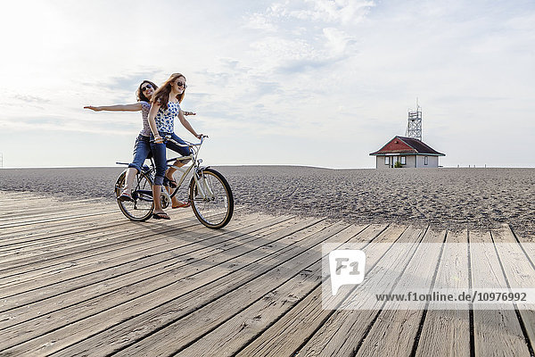 'Two girls riding double on a single bike on a beach boardwalk; Toronto  Ontario  Canada'
