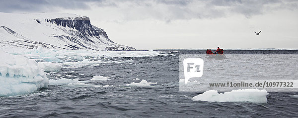 Menschen in einem Schlauchboot auf dem arktischen Ozean mit Blick auf die schneebedeckten Klippen entlang der Küste; Spitzbergen  Svalbard  Norwegen'.