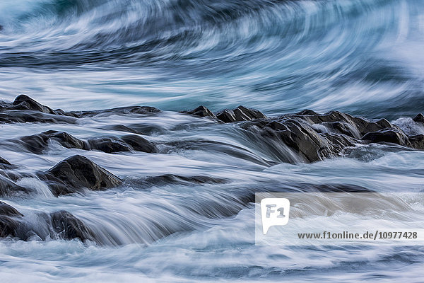 Langzeitbelichtung von Wellen  die auf die Küstenlinie treffen und über Felsen fließen; Island