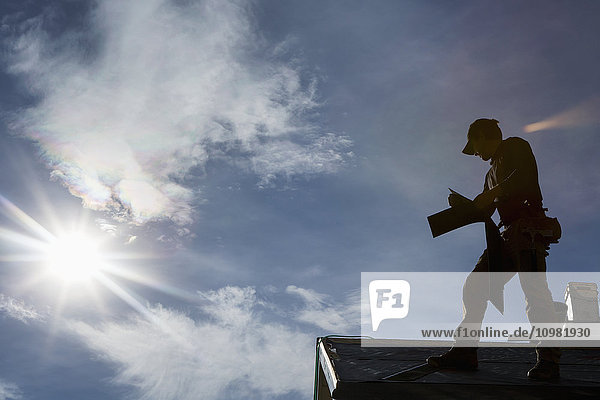 'Silhouette of male roofer on top of roof holding new shingles with clouds sun burst and blue sky; Calgary  Alberta  Canada'