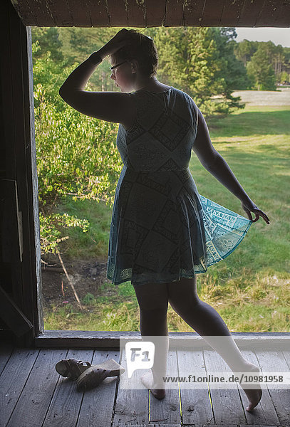 Ein rothaariges  weißes Teenager-Mädchen in einem türkisfarbenen Kleid und ohne Schuhe steht auf dem Heuboden einer alten Scheune  umrahmt von einem offenen Dachfenster mit Blick auf leere Grasfelder; Pittsboro  North Carolina  Vereinigte Staaten von Amerika'.