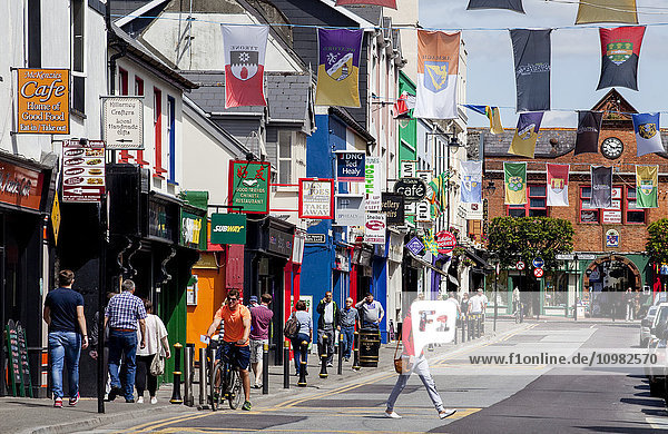 Fußgänger auf dem belebten Bürgersteig mit bunten Gebäuden und Bannern mit Wappen  die über der Straße hängen; Killarney  Grafschaft Kerry  Irland'.