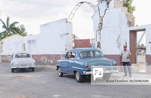 Eine Straßenansicht von Kuba mit Oldtimern  die neben heruntergekommenen Gebäuden geparkt sind  und einem Mann mittleren Alters  der auf dem Bürgersteig steht; Matanzas  Kuba'.