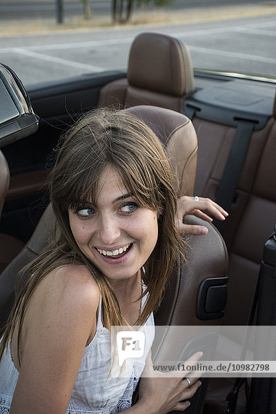 Lächelnde junge Frau sitzt im Cabrio und zieht lustige Gesichter.