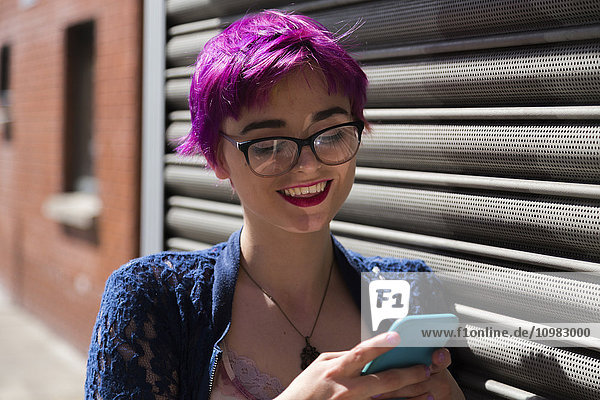 Porträt einer lächelnden jungen Frau mit gefärbtem Haar  die ihr Handy ansieht.