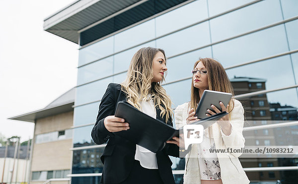 Zwei Geschäftsfrauen mit digitalem Tablett und Mappe vor dem Bürogebäude