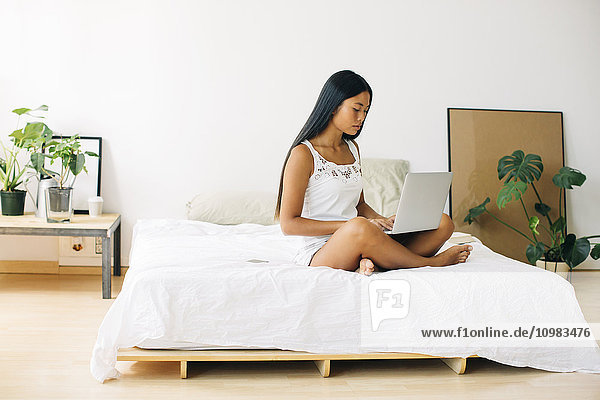Junge Frau auf dem Bett sitzend mit Laptop