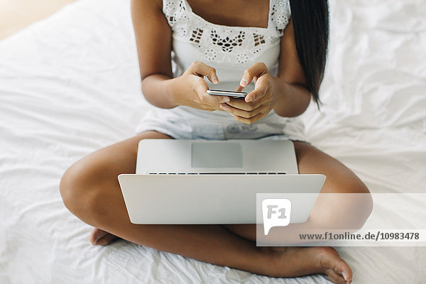 Young woman sitting on bed using cell phone and laptop