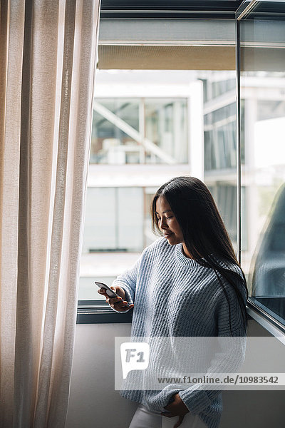 Young woman with cup of coffee standing in front of open window looking at cell phone