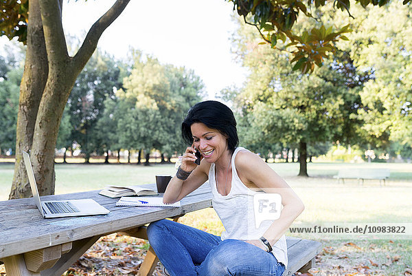 Lächelnde Frau sitzt auf einer Bank im Park und telefoniert mit einem Smartphone.