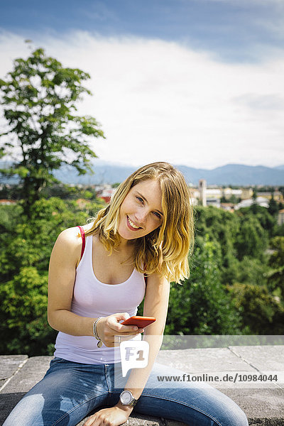 Lächelnde junge Frau mit Smartphone an der Wand sitzend