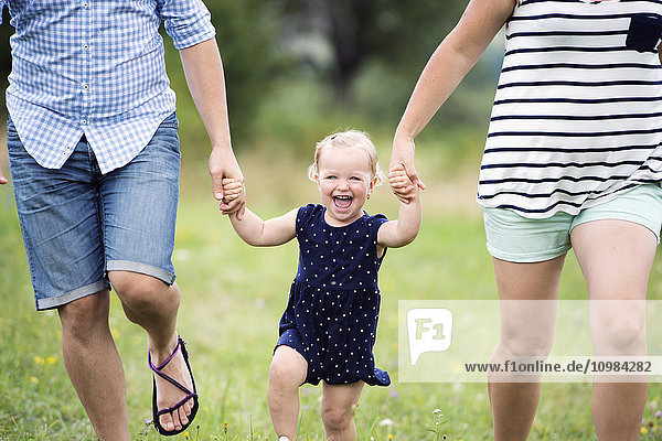 Lachendes Kleinkind Mädchen  das beim Laufen die Hände der Eltern hält.