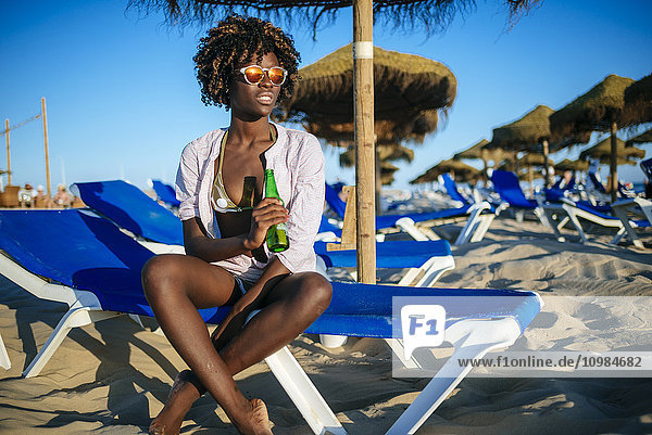 Junge Frau sitzt am Strand und trinkt Bier.