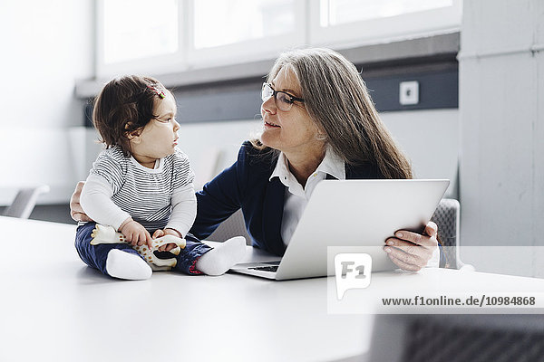 Senior Geschäftsfrau sitzend am Konferenztisch mit Laptop und Blick auf Baby Girl