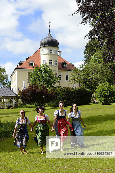 Deutschland  Bad Heilbrunn  vier glückliche Frauen mit Dirndln  die Hand in Hand auf einer Wiese laufen.