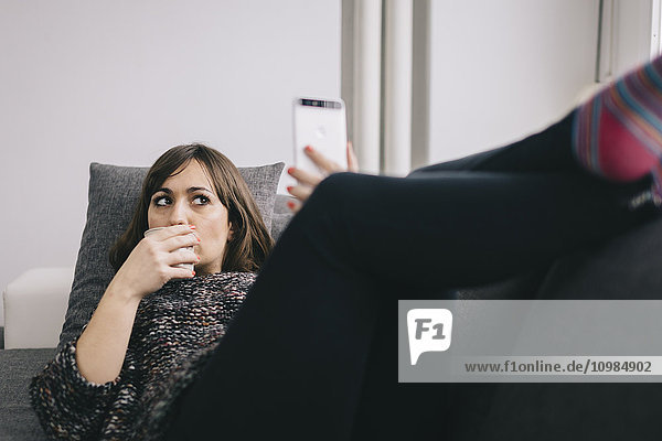 Junge Frau entspannt sich auf der Couch bei einer Tasse Kaffee und nimmt Selfie mit dem Smartphone.