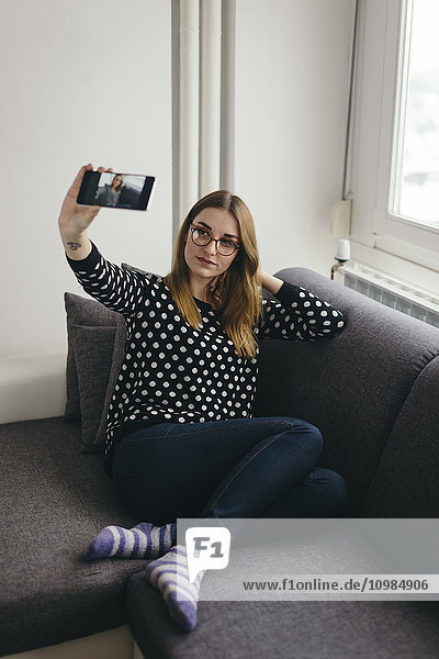 Junge Frau entspannt sich auf der Couch und nimmt Selfie mit dem Smartphone.