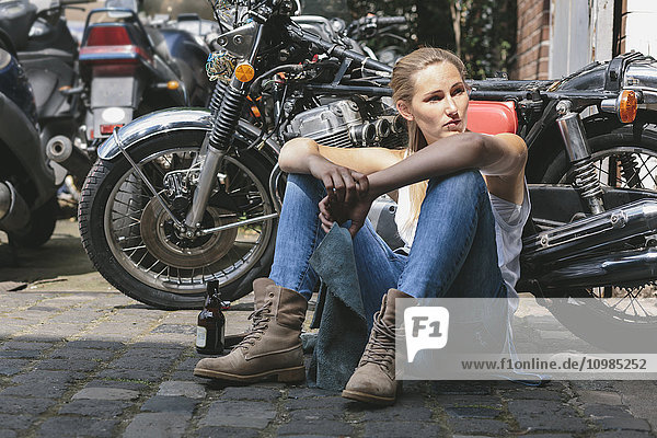 Young woman with beer bottle sitting next to motorbike