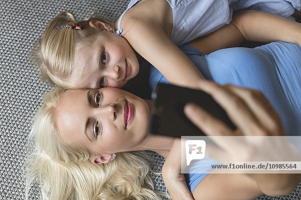 Mutter und ihre kleine Tochter liegen auf dem Boden und nehmen Selfie mit dem Smartphone.