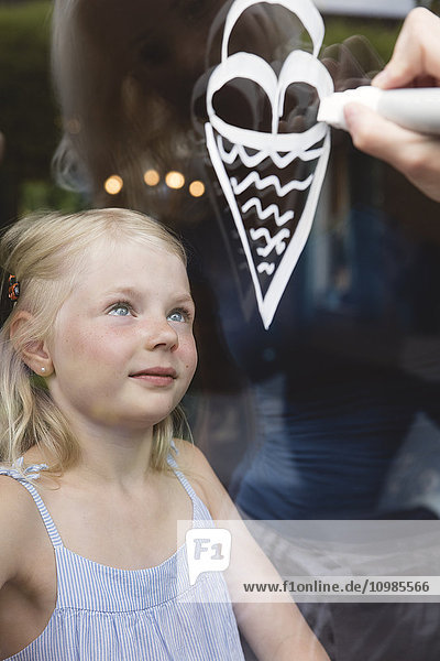 Porträt eines kleinen Mädchens hinter der Fensterscheibe  das seine Mutter beim Malen von Eiscremezapfen beobachtet.