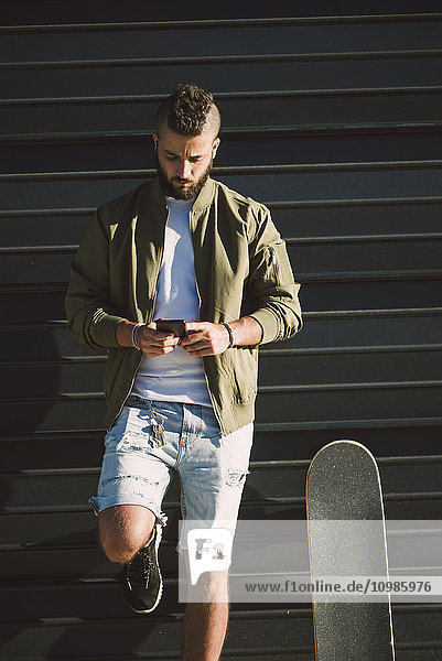 Mann mit Skateboard vor der Fassade mit Blick auf das Handy