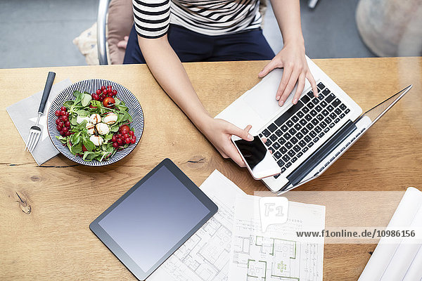 Woman at desk using laptop and cell phone next to construction plan and salad