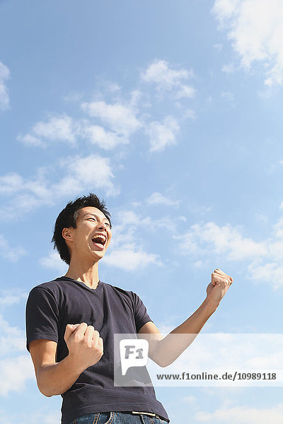 Young Japanese man cheering against blue sky