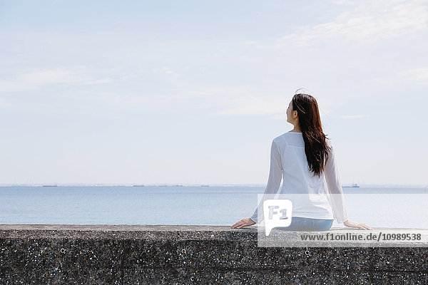 Junge japanische Frau sitzt auf einer Betonmauer am Meer