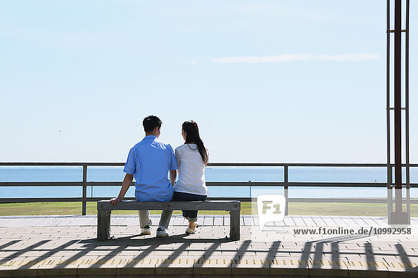 Junges japanisches Paar sitzt auf einer Bank am Meer