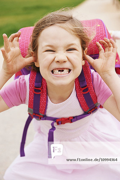 Porträt eines 5-jährigen Mädchens mit rosa und lila Schultasche  das Grimassen schneidet und in die Kamera blickt  wobei ihre Vorderzähne fehlen