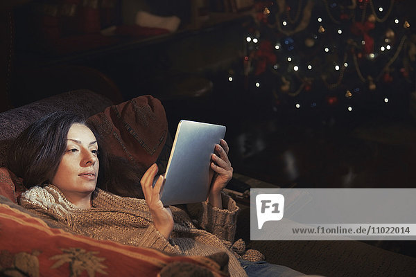 Frau mit digitalem Tablett entspannt auf dem Sofa im Wohnzimmer in der Nähe von Weihnachtsbaum