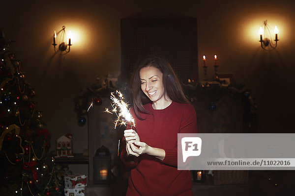 Lächelnde Frau mit funkelndem Feuerwerk im dunklen Wohnzimmer mit Weihnachtsbaum