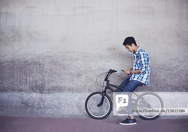 Teenage boy texting auf BMX Fahrrad an der Wand