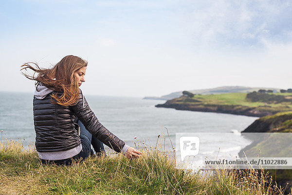 Rear view of woman sitting on hill by sea against sky Rear view of woman sitting on hill by sea against sky