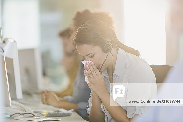 Geschäftsfrau mit Headset schnäuzt sich am Computer bei der Arbeit
