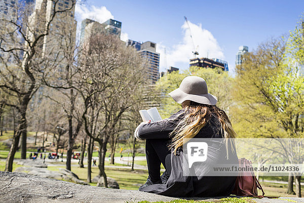 Rückansicht einer Frau die auf einem Felsen im Central Park sitzt und ein Buch liest