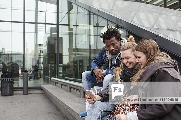 Friends looking at smart phone while sitting on retaining wall by glass building