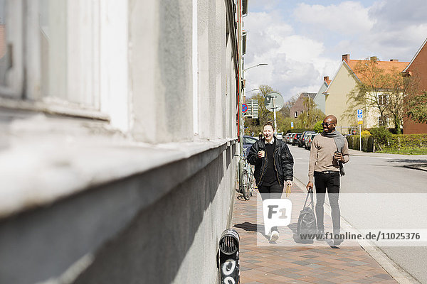 Happy friends holding coffee cups while walking on sidewalk during sunny day