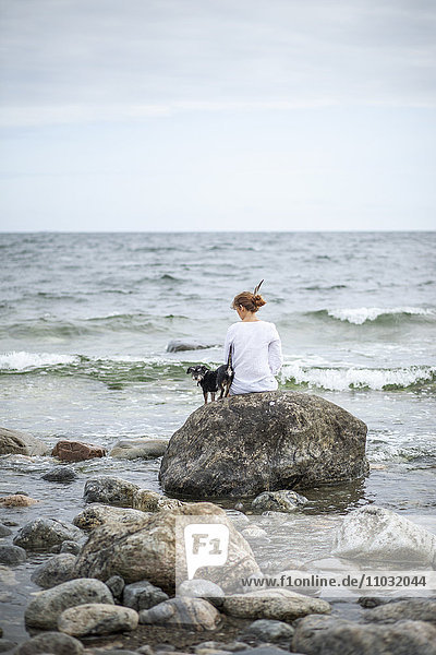 Frau mit Hund auf einem Stein am Meer sitzend