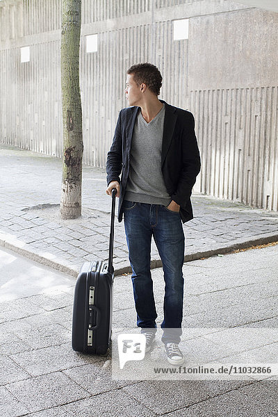 Junger Mann mit Koffer auf dem Gehweg stehend