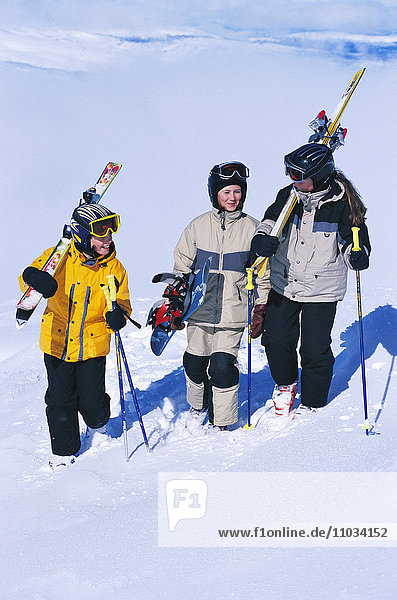 Three children trudging in snow carrying snowboard and slalom skis.