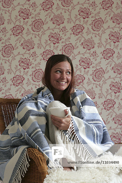 Scandinavian woman with a cup of coffee sitting with a blanket wrapped around her.