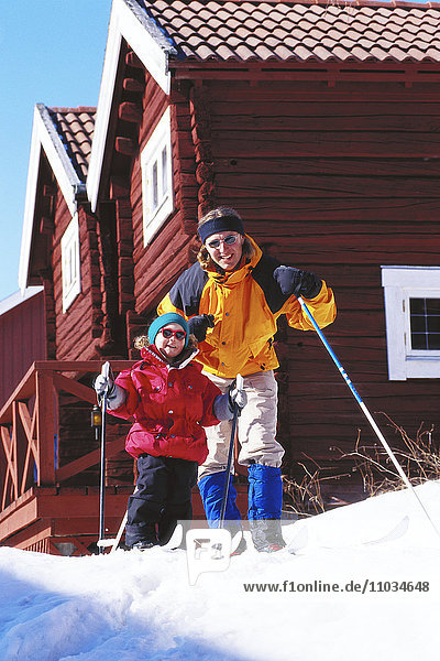 Eine Mutter und ein Kind auf Langlaufskiern vor einer Berghütte.