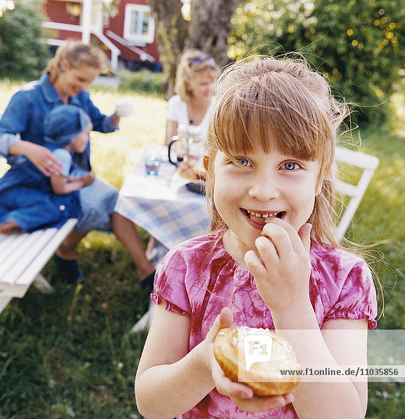 Ein lächelndes Mädchen  das zusammen mit anderen Kindern und Erwachsenen eine Kaffeepause im Garten macht.