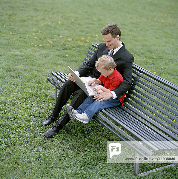 Geschäftsmann mit kleinem Jungen und Laptop auf einer Parkbank.