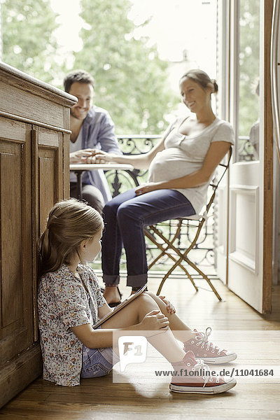 Mädchen auf dem Boden sitzend mit digitalem Tablett als Elternchat im Hintergrund