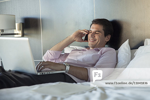 Man lying in bed  using cell phone and laptop computer