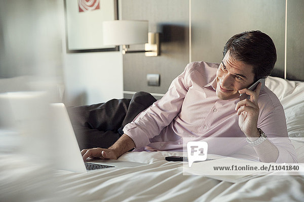 Man lying on hotel bed  talking on cell phone and using laptop computer